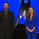 A screenshot from the 2020 DICE Awards depicting hosts Jessica Chobot and Greg Miller on stage against a very blue background.